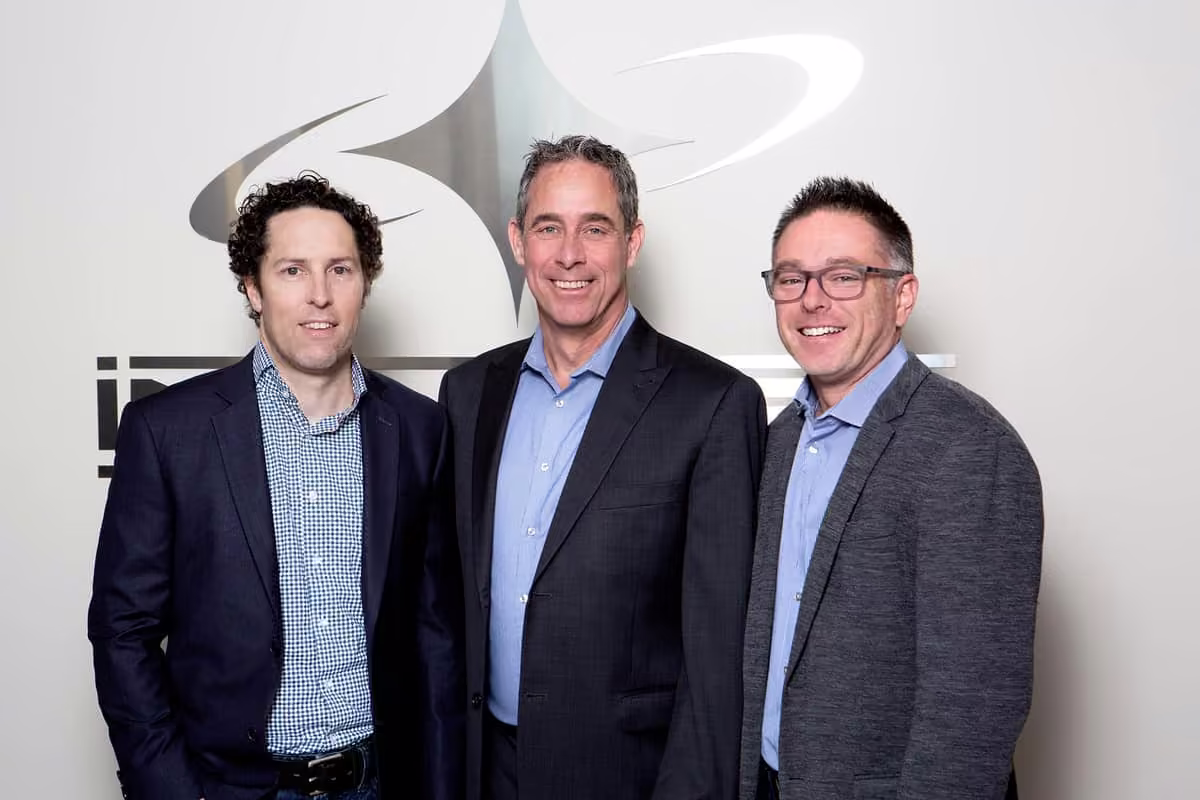 Trois hommes d'affaires souriants en vestons posant devant un mur avec un logo métallique